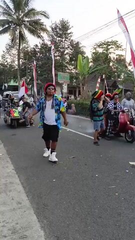 A man wearing a colorful floral shirt, white t-shirt, black shorts, white high-top sneakers, sunglasses, and a Rasta-style hat dances on a paved road. He has long, dark dreadlocks and wears a gold chain. He is moving his arms and legs as if dancing. Behind him, there are people on a multi-person vehicle that resembles a scooter or a small cart, adorned with Indonesian flags and Rasta colors. Other people are walking or standing on the side of the road, some also wearing Rasta hats. Indonesian flags are also displayed on poles along the roadside. The environment appears to be a rural or suburban area with trees and a building in the background. The sun is low in the sky, suggesting it is either early morning or late afternoon.