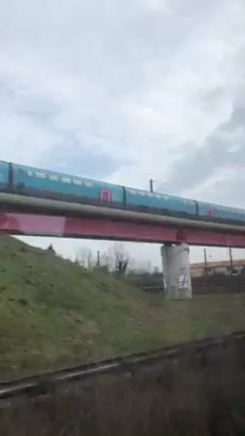The video shows a train moving across a bridge. The train is blue and red. The bridge is red. The landscape includes a grassy hill and a cloudy sky. Graffiti is visible on a retaining wall near the bridge. The view is from a moving vehicle.