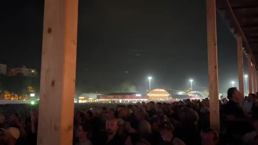 A nighttime outdoor concert is depicted. The camera is positioned behind several wooden pillars, looking out at a large crowd of people. In the distance, there are buildings with lights on, trees, and multiple stages or structures with bright lights and screens. Some people in the foreground are visible, and many are holding up their phones. The overall atmosphere suggests a lively event.
