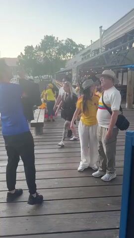 A group of people are walking on a wooden pier near a body of water. The person in the foreground on the right is a man wearing a white shirt, khaki shorts, and a hat. He is standing next to a woman wearing a yellow shirt and white pants. Both are looking towards the left. In the background, other people are walking and standing. Some are wearing yellow shirts with red, blue, and yellow stripes, suggesting they may be supporting a team or country. There is a building with outdoor seating to the right of the pier. The sky is visible in the background.