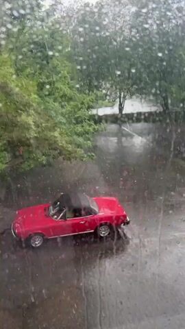 The video captures an elevated view of a red convertible car parked outdoors on a rainy day. The classic-style car, possibly a Fiat 124 Spider, features chrome accents and has its black soft top raised. The scene is viewed through a window, with numerous raindrops visible on the glass, creating a blurred effect over the landscape.

Heavy rain is actively falling, causing water to splash on the dark, wet paved ground, which appears to be a cobblestone or paver surface. The ground reflects the ambient light, indicating the wet conditions. In the background, lush green trees and other foliage are visible, suggesting a natural element within an urban or residential setting, possibly a parking area or driveway. The weather is clearly overcast and rainy, but it appears to be daytime. No people or visible text are present in the frame.