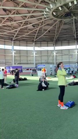 The video shows the interior of a large, indoor stadium-like building with a high, steel-beamed ceiling and large windows along the sides. The floor is covered in artificial green turf. There are numerous people scattered around, mostly sitting or lying down on the turf. Some people are walking around, and one woman is standing and talking on her cell phone. Many people have bags or backpacks near them. In the background, there are red banners with white lettering. The overall scene appears to be a resting or waiting area in a sports or event venue. The lighting is bright and natural.