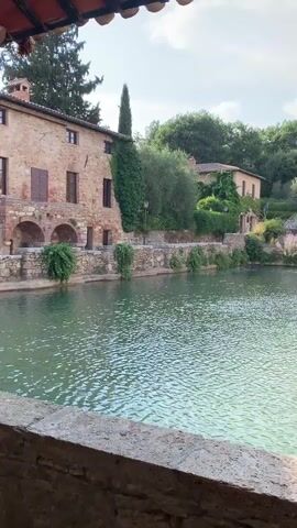 The video shows a wide shot of a natural thermal pool with turquoise water. The pool is surrounded by old stone buildings with red tile roofs and green shutters. Lush green trees and vegetation are visible in the background. The water is slightly rippled, suggesting a gentle breeze or the flow of thermal springs. The light suggests it is daytime, likely late morning or early afternoon, with some clouds in the sky. There are no people or animals visible in the frame. The camera pans slowly to the right, revealing more of the buildings and the pool's edge. The overall impression is one of tranquility and historical charm.