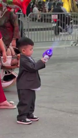 A young Asian toddler, dressed in a gray suit and black-and-white sneakers, is standing outside on a paved surface while holding a purple bubble gun that is emitting bubbles. The child is looking at the bubble gun. An adult wearing red pants and sandals is partially visible to the left of the child, along with a baby stroller and a red and green foil balloon in the shape of a fish. Several other people are visible in the background, along with a metal barricade. The environment appears to be a crowded public space.