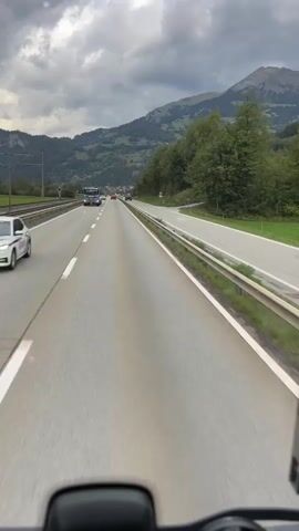 A first-person perspective video shows a highway under a cloudy sky. The camera is positioned in a vehicle, looking forward. The highway has multiple lanes, with a barrier on the right and a train track on the left. In the distance, there are mountains covered with trees. Several vehicles are visible on the highway, including trucks and cars. The video appears to be shot in motion, indicating the vehicle is driving.