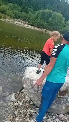 A young girl wearing a coral-colored shirt and black leggings stands on a large rock by a river, facing away from the camera. She holds her arms out in front of her. A person wearing a teal shirt and blue pants, with a black baseball cap, stands to her right, facing her and looking in the same direction. The person's right hand is extended, pointing at something in front of the girl. The river is calm and green, with trees lining the opposite bank. The riverbank in the foreground is covered in pebbles and rocks. The girl then appears to throw something into the river.