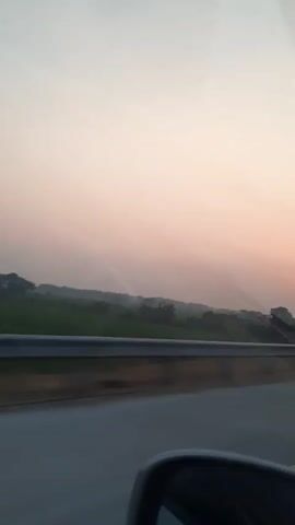 The video shows a view from a car window, likely during a sunrise or sunset. The sky is a pale pink and white gradient. In the distance, there are fields and trees. A road barrier is visible in the foreground, and part of the car's side mirror is in the lower right corner. The trees on the right side of the frame are blurred due to the car's motion. A faint rainbow-like arc is visible in the sky above the fields. The sun is a small, bright orb low on the horizon, partially obscured by trees.