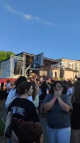 A group of young people are gathered outdoors in front of a stage. The stage is set up with speakers and lighting equipment. Some people are looking towards the stage, while others are talking amongst themselves. One person in the foreground is holding their hands to their face, appearing surprised or shocked. The sky is clear and blue, suggesting it is daytime. There are buildings in the background, indicating an urban or suburban setting.
