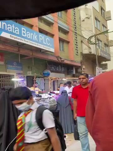 The video shows a crowded street scene. The camera's point of view is behind a person wearing a red shirt. In the background, there are several buildings, including a bank. A market scene is evident with vendors selling clothing and other goods.  There are people walking by, many wearing traditional Islamic clothing. Several yellow autorickshaws are parked along the street. Overhead are power lines and street lights.