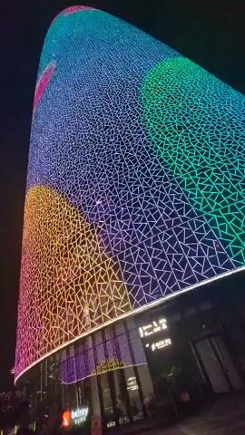 The video shows two tall, cylindrical buildings covered in a geometric light display at night. The lights on the buildings are a mix of vibrant colors, including pink, purple, blue, green, yellow, and orange, forming abstract patterns. The buildings are illuminated from below, with the light patterns wrapping around their curved surfaces. The camera is positioned at ground level, looking up at the buildings. In the foreground, there are some blurred figures of people walking, and the lower sections of the buildings appear to be commercial spaces with signs and glass windows. The sky is dark, indicating nighttime. The video is filmed from a low angle, emphasizing the height and grandeur of the illuminated structures. The overall impression is one of a modern, dynamic urban environment with striking architectural lighting.