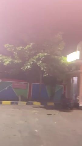 A night time video shows a paved area outside a building. The building appears to have a storefront with glass doors and windows and an illuminated sign. A white car is parked in front of the building, and a motorcycle is parked to the left. Trees and foliage are visible to the left of the building. The lighting is dim, and there is a slight haze in the air. The camera pans to the right. A person wearing a light-colored shirt and a hat rides a motorcycle away from the camera and toward the building. The video ends.