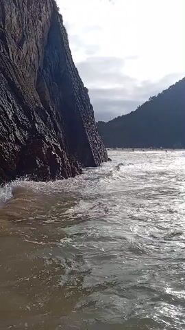 This media file captures a scenic coastal view in Ribadedeva, Spain. The scene features a rugged cliff face on the left, with waves crashing against its base. The water is a murky green, reflecting the light. In the distance, rolling hills covered in dark vegetation form a backdrop. The sky is partly cloudy, with patches of bright sunlight suggesting a day that is transitioning between overcast and clear conditions. The overall mood is one of natural beauty and the power of the ocean. No text is visible in the media file.