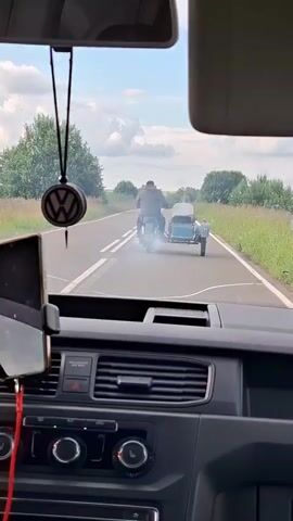 The video shows a view from inside a car driving on a paved road. The road is lined with tall green grass and trees, suggesting a rural or countryside setting. The sky is partly cloudy, indicating a bright but not entirely sunny day. In front of the car, a motorcycle with a sidecar is traveling in the same direction. The motorcycle appears to be emitting a white smoke or steam from its rear, possibly due to a mechanical issue or a deliberate effect. The rider is wearing a helmet and jacket. The interior of the car is visible in the foreground, with a dashboard, steering wheel, and a phone mounted on the windshield. A Volkswagen logo hanging from the rearview mirror is also visible. The scene appears calm and ordinary, except for the smoke from the motorcycle. The overall impression is one of a casual drive through the Russian countryside.