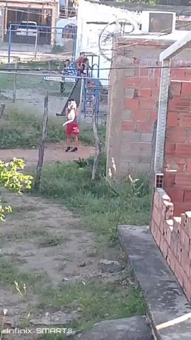A video shot from an elevated perspective shows a boy in red shorts and a white shirt standing outside a fenced area, holding a white object, potentially a ball. In the background, several people are visible on playground equipment. A partly constructed brick wall frames the right side of the shot, while a green, leafy bush is on the left. The background includes buildings and a bicycle mounted on a wall. A "Infinix Smart 8" watermark is visible at the bottom left corner of the screen. The ground is covered in patches of grass and dirt.