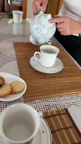 A person's hands are seen pouring tea from a white teapot with a delicate floral pattern into a white teacup, which sits on a matching saucer with a blue rim and floral accents, all placed on a bamboo placemat. The table is covered with a white, patterned tablecloth. Alongside the teacup, an oval white plate holds various cookies, some appearing to be shortbread with sesame seeds, and others with an oatmeal or raisin texture. A small round white plate next to it contains four small candies or chocolates, two brown and two pink, each in a white paper cup. Another empty teacup is visible in the foreground, and a blurred background shows parts of a room, possibly a dining area or kitchen. The activity suggests a tea-making and serving moment, with freshly brewed tea being poured.