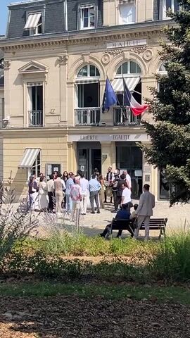 A group of people are gathered in front of a building with "MAIRIE" written above the entrance. Two flags, one blue and one red, white, and blue, are displayed from the building. The words "LIBERTE EGALITE" are visible below the flags. Some people are standing and talking, while others are seated on a bench in the foreground. There is lush greenery in the foreground and to the right of the building. The weather appears to be sunny.