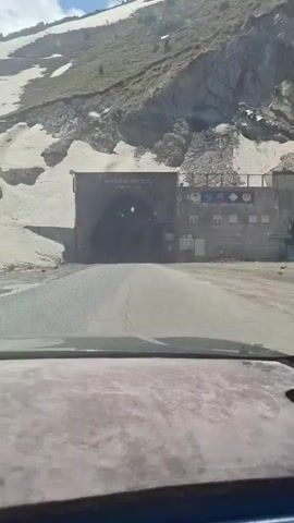 The video shows a car driving towards a tunnel. The tunnel is labeled "NAKI ISTIKLOL" in both Cyrillic and Arabic script. The tunnel entrance is a dark, gray block of concrete. There is a building to the right of the tunnel entrance, with a series of windows. The road leading to the tunnel is paved and gray. There is snow visible on the rocky hills surrounding the tunnel. The camera is inside the car, showing the view from the driver's seat. The dashboard is visible in the foreground. As the car gets closer, the tunnel lights appear, and then the car enters the tunnel, making the video completely dark.