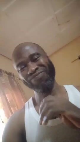 A man with a bald head and a beard, wearing a white tank top, rubs his neck and chin. He smiles slightly and looks at the camera. The background includes a patterned curtain and a white ceiling with a grid pattern. The video appears to be shot from a low angle.