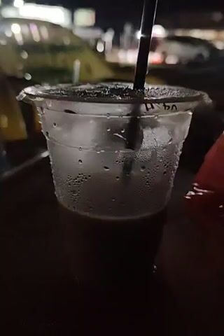 The video depicts a close-up of a plastic cup with a black straw, containing a dark liquid, possibly coffee. Condensation is visible on the exterior of the cup. The camera then pans out, showing a table with two plastic chairs, also with condensation. In the background, there's a view of an outdoor area at night, possibly a beach or park, with lights and figures of people and motorcycles visible. The scene suggests an evening setting, perhaps at a casual outdoor cafe.