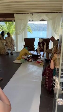 A long-shot video captures a traditional Indian wedding ceremony taking place indoors with a picturesque lake view in the background. The venue features large glass windows draped with white curtains, allowing ample natural light into the space. A white runner stretches across the dark tiled floor, leading to a central area where a man in a yellow traditional outfit kneels, preparing items on the floor. A wooden structure is in front of him. The room is populated with wedding guests, many dressed in traditional Indian attire, seated on white chairs. 

A couple sits towards the back of the room, also in traditional clothing. Seated on either side of the man in yellow are two men in western-style clothing. One of them wears a turban. The camera pans slightly, revealing more guests seated throughout the room. The overall atmosphere appears calm and reverent as the preparations for the ceremony proceed.