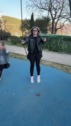 A woman in a black outfit and pink-striped sneakers swings on a swing set in a park. She is wearing a black leather jacket, black pants, and a black shirt, along with pink tinted sunglasses. The swing set has a blue rubber ground. The woman is smiling and swinging back and forth, pushing off the ground with her feet. There are trees and a hill in the background.