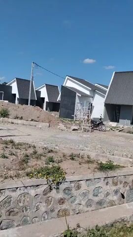 This is a video of a residential construction site under a clear blue sky. Several houses, in various stages of completion, are visible. They are designed with modern, angular roofs and dark gray siding. Construction materials, such as scaffolding, bricks, and wheelbarrows, are scattered around the properties. A motorcycle is parked in front of one of the houses. The ground is covered with dirt and gravel, and there is a concrete drainage ditch in the foreground. The overall impression is of an active construction zone.