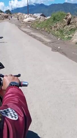 The video shows a first-person perspective of someone riding a motorcycle on a dirt road. In the foreground, the rider's hands are visible on the handlebars, and a watch is on their left wrist. The rider is wearing a maroon jacket with a checkered patch on the sleeve. The road is unpaved and appears rough, with loose gravel and dirt. To the right of the road, there is a steep embankment covered in vegetation and large rocks. To the left of the road, there are more rocks and debris, and in the distance, a body of water, possibly a lake, is visible. Mountains can be seen in the background under a clear blue sky with scattered white clouds. Several other motorcycles are visible on the road ahead, some with riders. One motorcycle approaches the camera, ridden by a person wearing a helmet and a jacket. The overall scene depicts a scenic but rugged outdoor environment, likely in a mountainous or hilly region, with the activity of motorcycle riding being central to the footage.