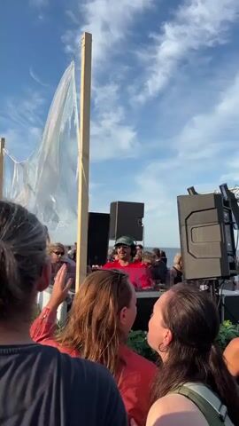 This is a vertical, eye-level video shot from behind a crowd of people looking towards a DJ who is performing outdoors, possibly at a beach or waterfront location given the sky and distant water visible. The DJ, wearing a red shirt and a hat, is standing behind a console with two large speakers on either side. He is actively engaging with the crowd, moving his arms and hands. Several people in the foreground are partially visible, with their backs to the camera, facing the DJ. Some are raising their hands, indicating they are enjoying the music. In the background, more people are gathered, and a white fabric decoration is suspended from a wooden pole. The sky is blue with scattered white clouds. The overall atmosphere appears to be a lively outdoor event.