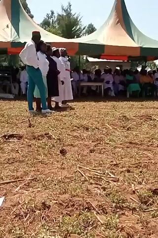 The video depicts a group of people standing in front of a seated audience under a tent. There are several people standing in the front, including one wearing a red hat and blue pants. Some are wearing white gowns. The audience under the tent is composed of many people of different ages. The ground is covered with dried grass and some green patches. The sky is clear and blue.