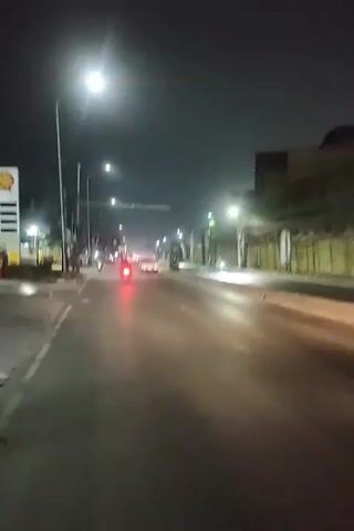 The video shows a view from a vehicle at night, moving down a city street. Streetlights illuminate the road and sidewalk. There are other vehicles on the road, including motorcycles and cars. Some of the vehicles have their headlights and taillights on. There are buildings and trees visible along the side of the road. The sky is dark.