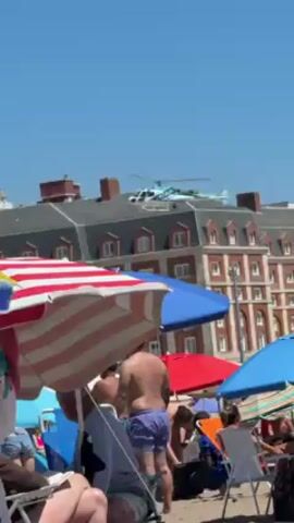 The video shows a crowded beach on a sunny day, with numerous beach umbrellas providing shade. A helicopter with a distinctive blue and white striped pattern flies at a low altitude in the background, behind a large building with a grey roof and brick facade. The building also features several chimneys. 

The foreground is filled with beachgoers, some sitting in chairs under the umbrellas. People are wearing beach attire such as swimsuits and shorts. There is a mix of individuals and groups, and some seem to be talking.