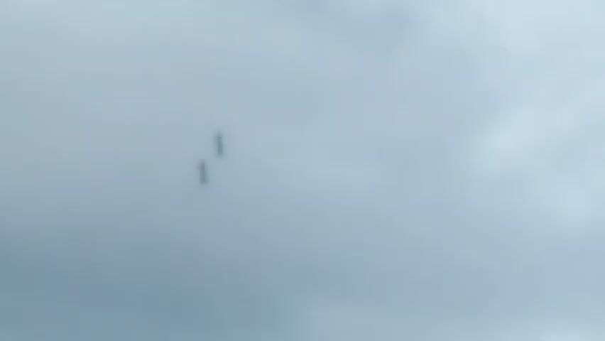 The video shows two small, dark aircraft flying in formation against a cloudy, light gray sky. The aircraft appear to be performing some kind of aerial maneuver, as one of them is emitting a thin trail of orange smoke. The camera is positioned below the aircraft, looking upwards. The image quality is somewhat blurry, making it difficult to discern specific details about the aircraft or their environment. The timestamp at the beginning indicates the video starts at 00:00 and ends at 00:04.