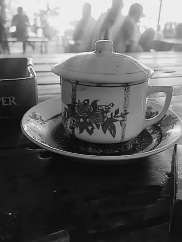 A black and white video shows a close-up of a teacup and saucer on a wooden table. The teacup has a lid and is decorated with floral patterns. To the left of the teacup is an ashtray with the words "ER" visible. In the background, several people are blurred and out of focus, suggesting they are in a public space such as a cafe or market. The overall lighting is bright, with a hazy quality that might indicate a sunny day or the presence of steam from the teacup. The focus is sharp on the teacup and saucer, while the background is soft. There are no discernible actions or interactions captured in the video; it appears to be a still shot with a slight zoom or panning movement. The environment seems to be outdoors or in an open-air setting due to the natural light and the presence of other people in the background.