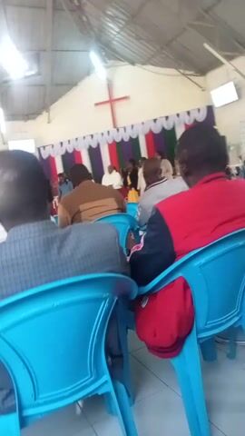 The video shows a group of people sitting in bright blue plastic chairs inside what appears to be a church. They are facing forward, most with their heads bowed, likely in prayer or contemplation. A cross is visible on the wall behind them. The building has a high ceiling and appears to be of simple construction. The camera angle is slightly off-kilter, giving a tilted perspective of the scene.