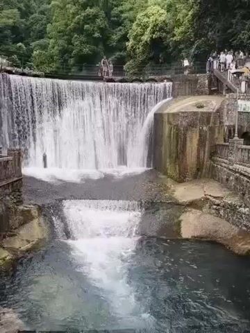 The video shows a tiered waterfall with a pool of water at the bottom. There are several people standing on a walkway and a set of stairs next to the waterfall. The area is surrounded by trees and lush greenery. The water is flowing down the tiers of the waterfall and creating a misty spray. Some people appear to be taking pictures or observing the waterfall. The scene is a natural landscape with some man-made structures for viewing.