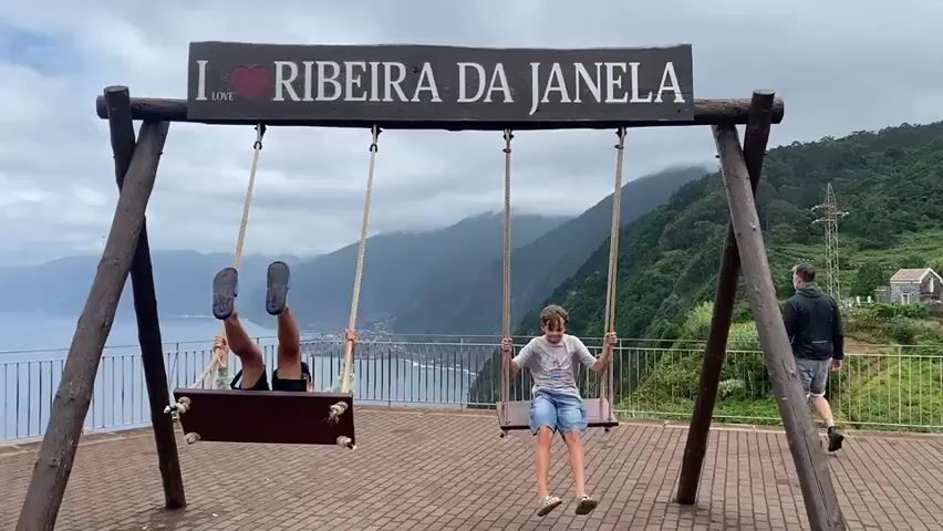 A wide shot shows a wooden swing set with two boys on swings, against a backdrop of mountains and the ocean. The swing set has a sign above it that reads "I LOVE RIBEIRA DA JANELA". The boy on the left swing is wearing a blue baseball cap, a green hoodie, and dark shorts. The boy on the right swing is wearing a white t-shirt and blue shorts. Both boys are swinging back and forth. The ocean is visible to the left, with steep, vegetated mountains in the background. The sky is overcast. To the right of the swing set, a person wearing a backpack is walking away from the camera. The ground around the swing set is paved with red bricks.