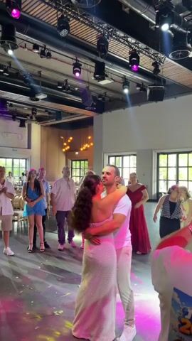 A wedding reception is taking place in a large indoor venue. The bride and groom are dancing closely in the center of the dance floor, bathed in colorful stage lighting. Several guests are gathered around, observing the couple, with some dancing in the background. The environment appears to be decorated with a modern aesthetic, featuring exposed ceiling beams and industrial-style lighting fixtures. The overall mood is festive and celebratory.