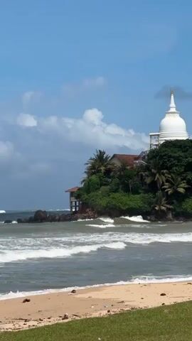 A vertical shot captures a serene beach with gentle waves rolling onto the sand. In the background, a lush green island juts out from the ocean, crowned by a white Buddhist stupa. The island is densely covered with tropical trees, including palm trees, creating a vibrant contrast with the blue sky dotted with fluffy white clouds. A traditional-looking building with a red-tiled roof is nestled amongst the trees on the island, with a small structure supported by stilts extending over the water. The sand in the foreground is light brown, with some scattered debris. The overall scene evokes a sense of tranquility and natural beauty.