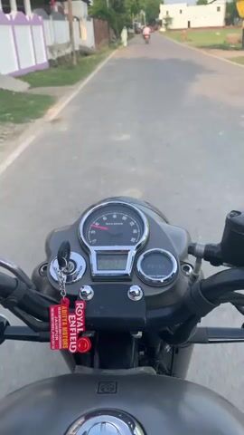 The video begins with a close-up of a motorcycle's handlebars and instrument cluster, featuring a speedometer, an LCD screen, and a fuel gauge. The camera then pans out to reveal a paved road with a shoulder on the left side. Trees and buildings line the road. In the distance, a person is walking, and another person is riding a motorcycle. The sky is visible and appears to be a light blue with some clouds. The overall scene suggests an outdoor environment, possibly a rural or suburban area, with mild weather conditions.