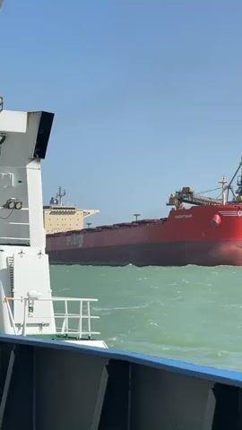 A large red bulk carrier, named "KNIGHTSHIP" and bearing "SeaEnergy" on its side, is seen docked or maneuvering near an industrial port under a clear blue sky. Water is being sprayed from the bow of the red ship. The video is filmed from a smaller white boat that is moving, providing a panoramic view of the port facilities.

The water is a choppy, greenish-blue, suggesting windy conditions. On the shoreline, various industrial structures are visible, including large conveyor belts extending over the water, buildings, and silos, indicating a busy shipping and industrial area. The overall scene depicts a functional port environment on a bright, sunny day.
