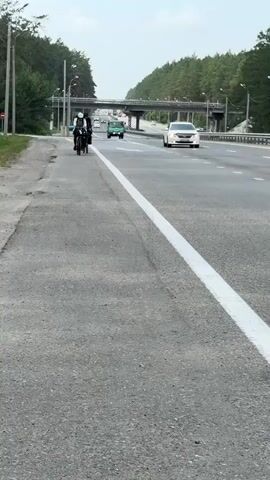 A motorcycle with two riders passes by on a paved path next to a highway. The rider in front wears a white helmet and a black jacket. The rider behind wears a black helmet and a black jacket. The motorcycle is dark colored. A green truck and a white car are driving on the highway. Trees line the sides of the highway and a bridge is visible in the distance. The sky is overcast.