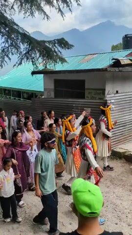 A group of people are gathered outdoors, possibly in a village setting, with a mountainous backdrop. Several individuals in traditional attire, characterized by elaborate headdresses with yellow and orange tassels and plaid sashes, are performing a dance or ritual. They wear long, white skirts and red-trimmed vests. Spectators, including women, children, and men, are watching the performance, some capturing the event with their phones. The environment features rustic buildings with corrugated metal siding and tiled roofs, surrounded by trees and vegetation. The ground is a mix of dirt and pavement.