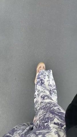 The video begins with a close-up shot of a person's legs and feet, wearing patterned pants and light-colored shoes, standing on a gray surface. The camera then pans up and moves to follow a man in a black shirt and pants walking down an airport jet bridge. He is pulling a black rolling suitcase behind him. The jet bridge has large glass windows on the right side, offering a view of the tarmac, an airplane with a purple tail, and the sky. The airplane appears to be parked and awaiting passengers or departure. Other passengers can be seen in the distance walking through the jet bridge. The lighting suggests it is daytime.