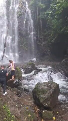 The media file captures a beautiful natural scene featuring a multi-tiered waterfall cascading down a rocky cliff face. The setting is lush and tropical, with dense green foliage surrounding the waterfall and the stream below. The water is a bright white as it plunges, creating a misty atmosphere around the base.

In the foreground, there are two individuals standing on the rocks near the stream. One is a person wearing a black t-shirt and black pants, standing with their back to the camera, seemingly observing the waterfall. Beside them, a younger person, possibly a child, is also standing on a rock, wearing a pink and yellow outfit and a backpack. They appear to be interacting or posing for a photo, with the younger person looking towards the waterfall. The ground is a mixture of dirt and rocks, with some moss and fallen leaves visible. A large, dark-colored boulder is prominent in the lower right corner of the frame.

The overall impression is one of a serene and picturesque natural landscape, likely a tourist destination in a tropical region like Banjar, Indonesia. The lighting suggests it is daytime, possibly overcast given the diffused light and the slight mist in the air. The activity taking place is simply enjoying the scenery and possibly taking in the experience of being at the waterfall. There is no visible text in the media file.