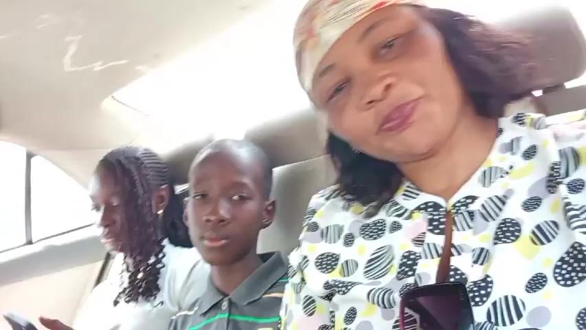 A woman with dark hair and a headscarf is in the back seat of a car, recording a video. Two children, a girl with braided hair and a boy, are seated beside her. The girl is looking at her phone. The woman is talking. The scene is brightly lit.