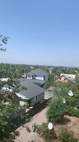 The video shows a view from a high vantage point overlooking a neighborhood with several houses and trees. The sky is clear and blue. In the foreground, green leaves and branches of a tree partially obscure the view. Below, a dirt path runs alongside a fence. Several houses with pitched roofs are visible, some with satellite dishes. The houses appear to be of various sizes and architectural styles. The overall scene depicts a residential area on a sunny day.