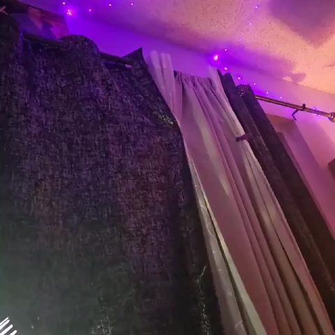 The video shows a room decorated with purple string lights along the ceiling. The camera is positioned low, looking upwards towards a window covered with gray and white curtains. A blue flame appears in the foreground, emitting from a torch-like device. The flame flickers and fades before disappearing, leaving the curtains as the sole focus of the view.