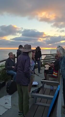 A group of people stand on a paved outdoor area overlooking the ocean at sunset. Several people are wearing hats. One person in the foreground is wearing a denim jacket with a large embroidered Buddha face on the back. There are musical instruments visible, including a guitar case and possibly a keyboard. Some people are sitting on benches. The sky is filled with clouds illuminated by the setting sun.