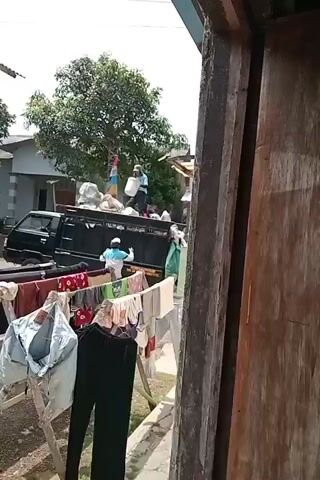 In an outdoor setting, two individuals are visible interacting with a black pickup truck. The truck is loaded with various items, including sacks and what appear to be bags. One person, wearing a blue shirt and a light-colored cap, is standing on the truck, holding a white bucket and seemingly transferring items. Another person, in a light-colored shirt and a cap, is positioned beside the truck, also handling items. In the foreground, a clothesline displays a variety of laundry, including shirts, pants, and undergarments, drying in the sun. The environment appears to be residential, with houses and trees in the background. The overall scene suggests a process of loading or unloading goods from the truck.