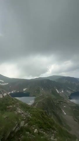 The video pans across a mountainous landscape featuring several serene lakes nestled among green hills under a cloudy sky. The camera starts from a higher vantage point, showing a wide view of the lakes and surrounding terrain. As the video progresses, the camera moves downwards, revealing more details of the mountainous terrain, including rocky slopes and patches of snow. A wooden railing is visible along the edge of the path, guiding hikers. In the latter part of the video, a few people are seen in the distance, some walking along the path, and others sitting near the edge, enjoying the view. The environment appears to be a popular hiking destination, with clear trails and well-maintained safety features.